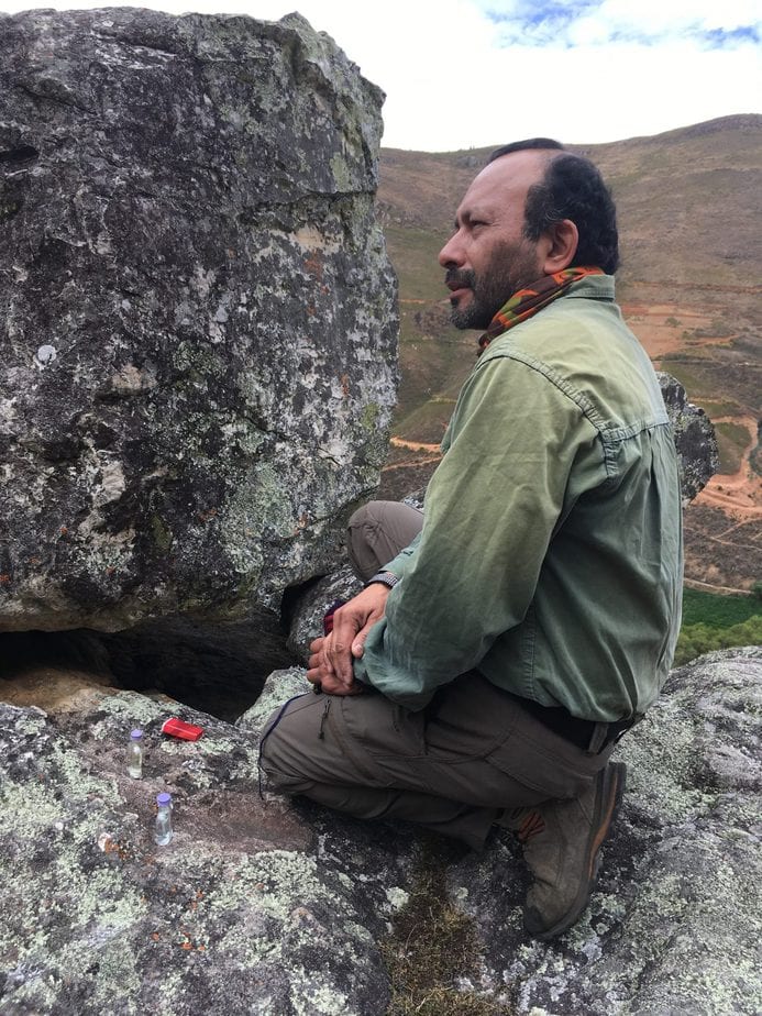 man facing left kneels in front of stones where he is making an offering