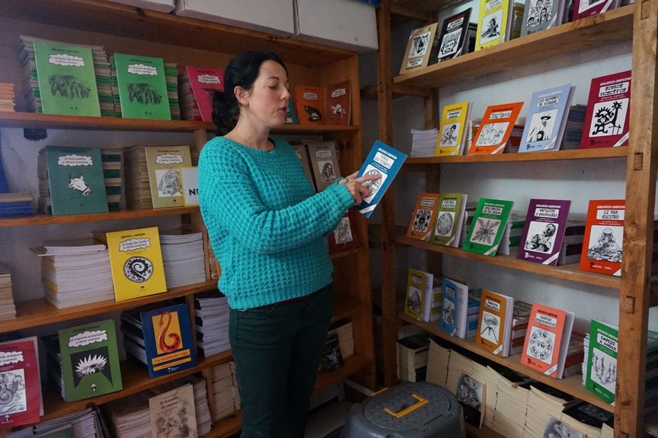 a woman holds a book in her hand and points at its cover. She stands in front of bookstands with the covers facing out.