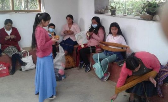 Women sit on a bench against the wall, knitting while a girl in a long skirt stands in the middle of the room