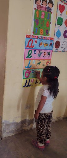 a little girl stands, back to the camera, pointing to a colourful posters showing letters of the alphabet