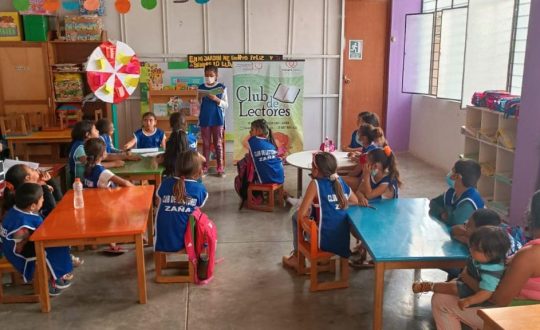 children in blue pinnies that say "Reding Club" sit at tables in a room while one girl stands at the front and reads