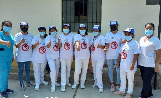row of people in white with masks give a thumbs up to the camedra. T-shirts say stop dengue.