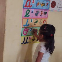 small girl, back to the camerca, points to a letter of a poster of vowels in script on the wall