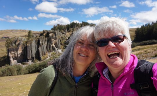two women, laughing, face camera, in the background a large rock formation against a cloudy blue sky