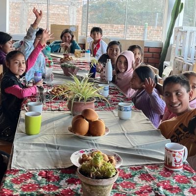 children seated around table raise their hands to wave at the camera