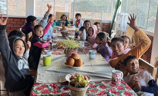 children seated around table raise their hands to wave at the camera