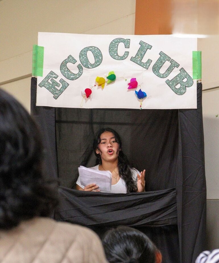woman speaks from behind a black stage made for puppets; a sign above her head reads "Eco Club"