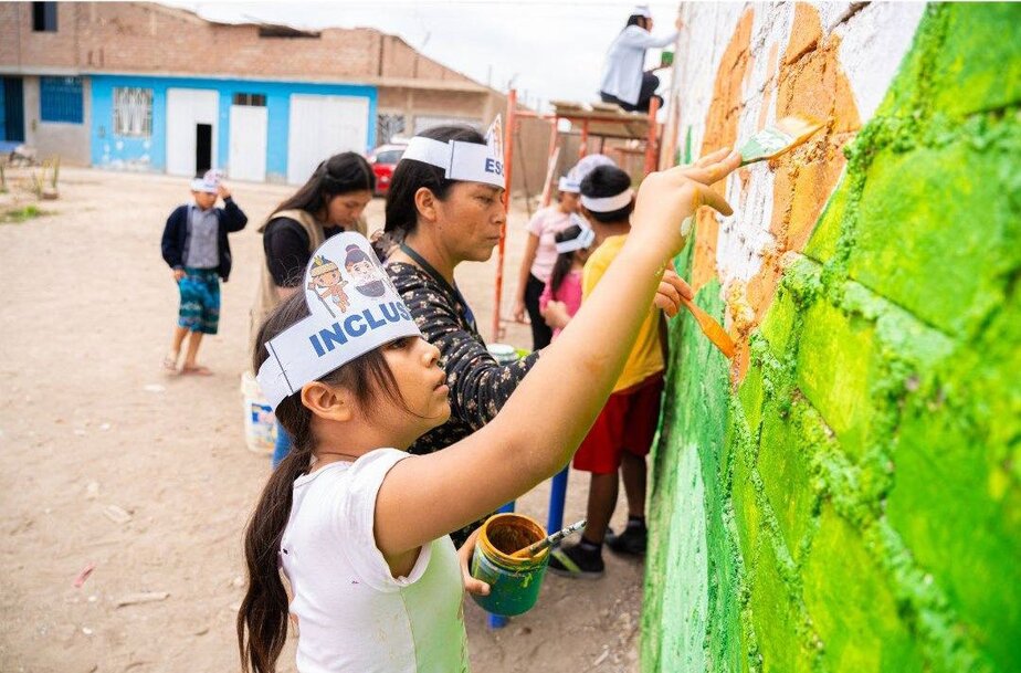 children and adults paint a mural on an outdoor cement block wall