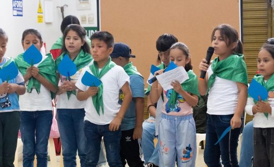 children wearing green kerchiefs stand side by side. One reads from a piece of paper into a microphhone.