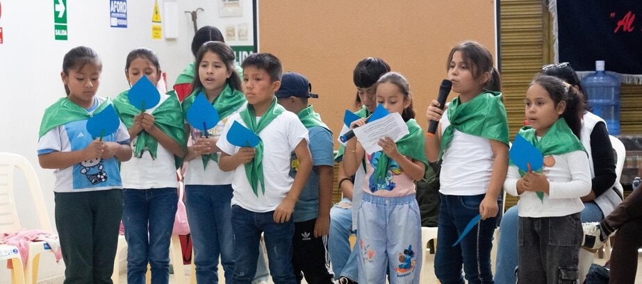 children wearing green kerchiefs stand side by side. One reads from a piece of paper into a microphhone.